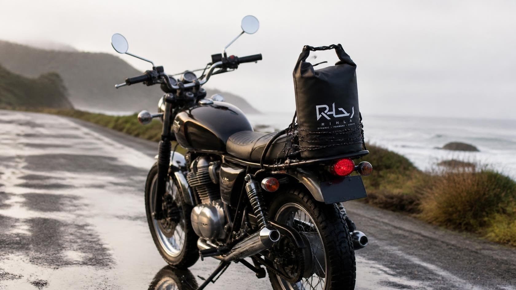 Black motorcycle on a wet coastal road with a black roll-top dry bag strapped to the rear